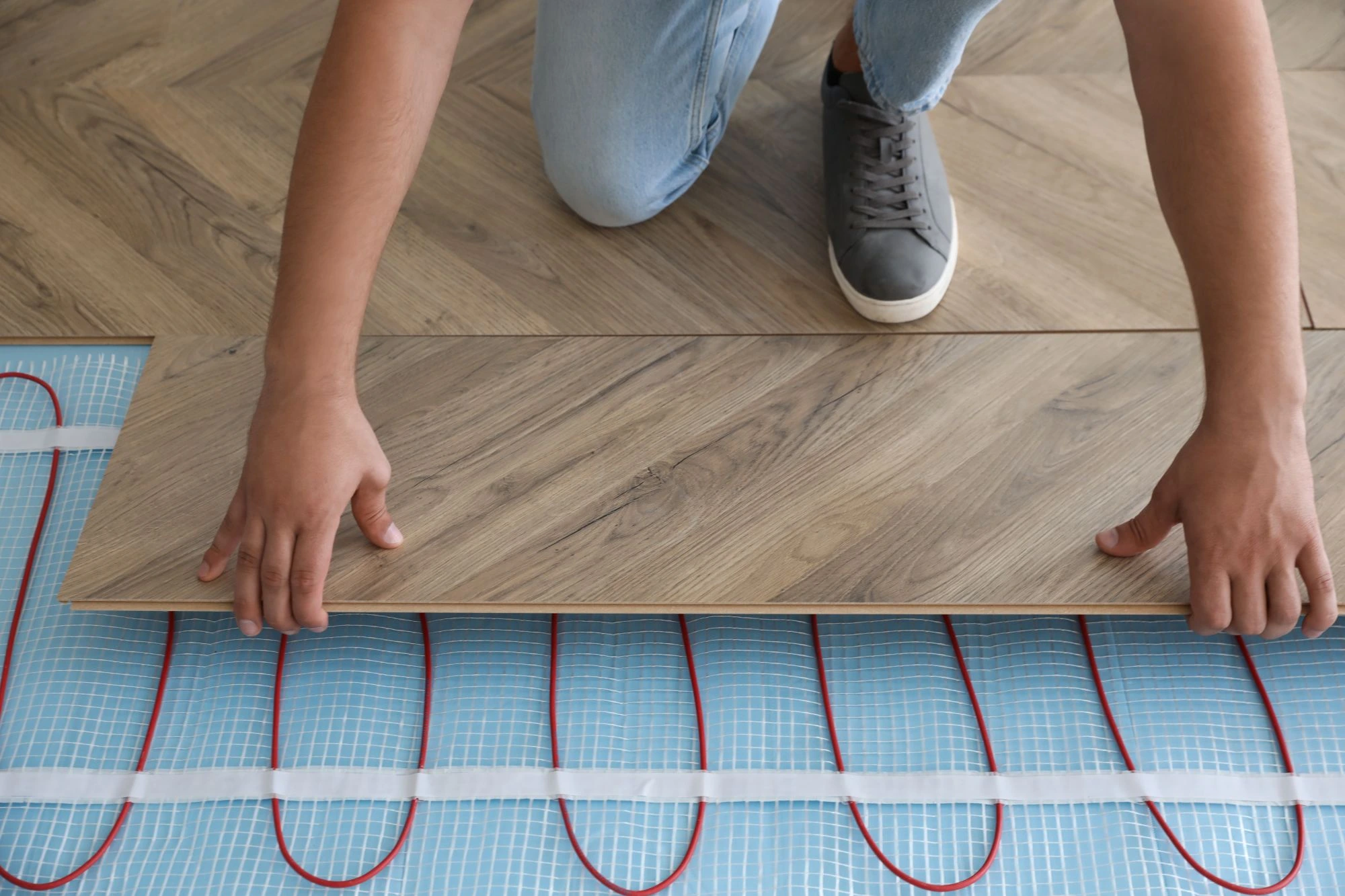 A person kneeling on a light wood floor, installing a wood-look laminate plank over a blue electric underfloor heating mat with red heating cables. 