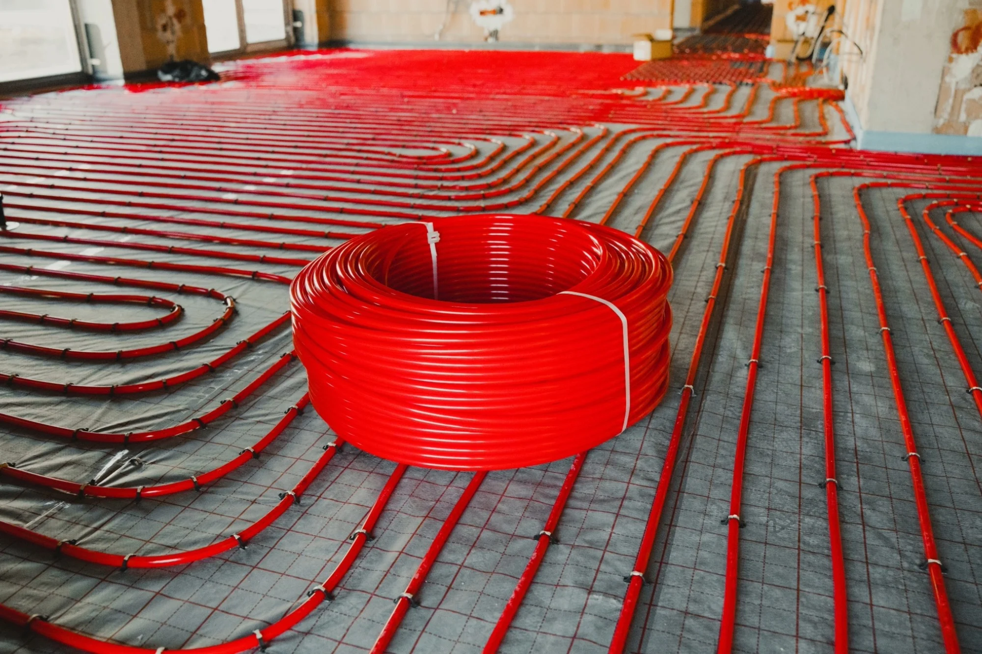 A large spool of red piping sits in the middle of a floor in the middle of a water underfloor heating system installation.
